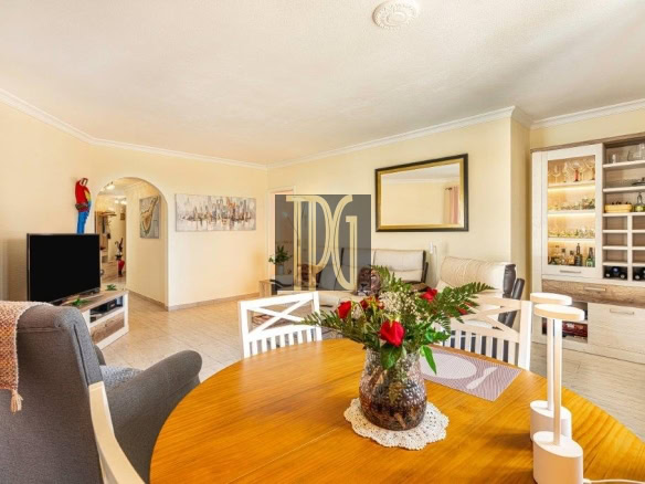Bright open-plan living and dining room with a round wooden table centerpiece of red flowers in a vase, white chairs, and a glass-front cabinet displaying bottles.