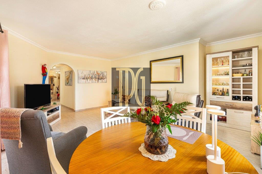 Bright open-plan living and dining room with a round wooden table centerpiece of red flowers in a vase, white chairs, and a glass-front cabinet displaying bottles.