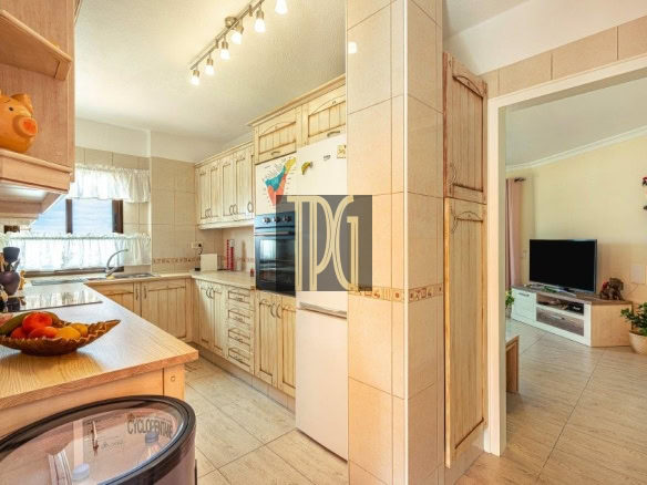 Bright kitchen with light wood cabinets, a fruit bowl on the island, and an open doorway to a living room with a TV and a decorative parrot wall ornament.