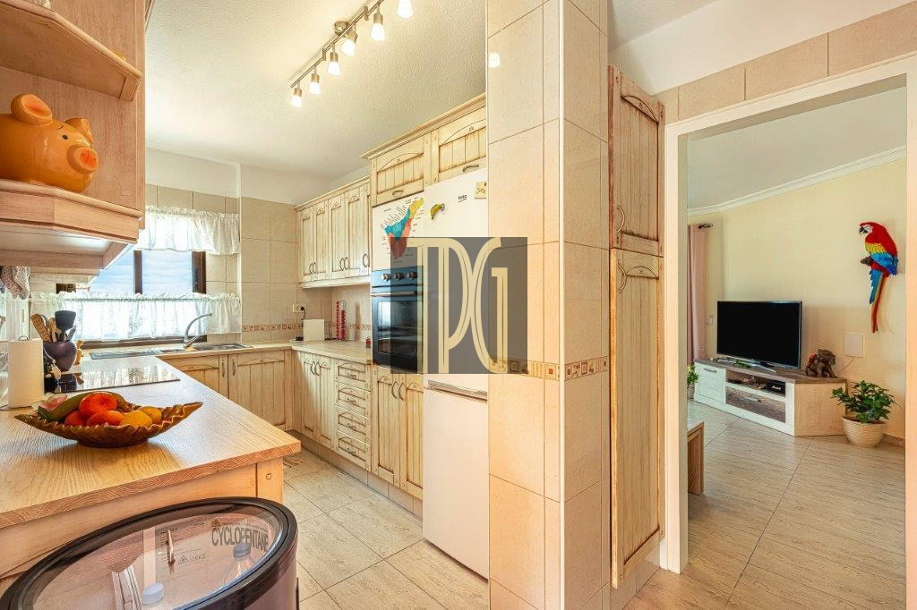 Bright kitchen with light wood cabinets, a fruit bowl on the island, and an open doorway to a living room with a TV and a decorative parrot wall ornament.