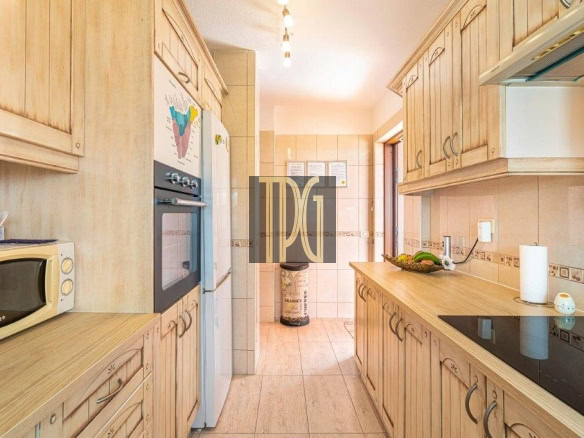 Bright kitchen with light wood cabinets, microwave, and built-in oven along a narrow galley wall. A long counter runs with a fruit bowl and paper towel holder on the right.