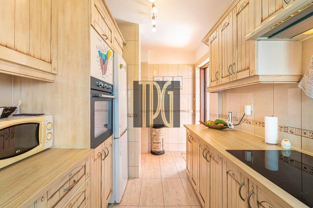 Bright kitchen with light wood cabinets, microwave, and built-in oven along a narrow galley wall. A long counter runs with a fruit bowl and paper towel holder on the right.