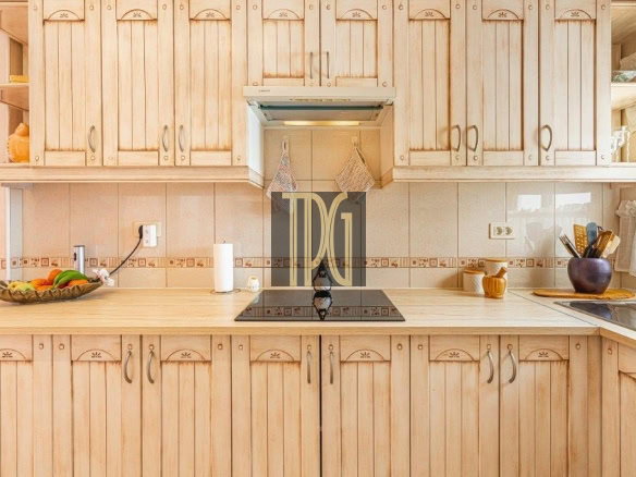 Sunlit kitchen with light wood cabinets, a black induction cooktop on a beige countertop, fruit bowl on the left.