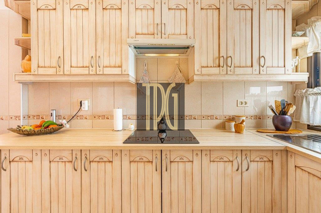 Sunlit kitchen with light wood cabinets, a black induction cooktop on a beige countertop, fruit bowl on the left.