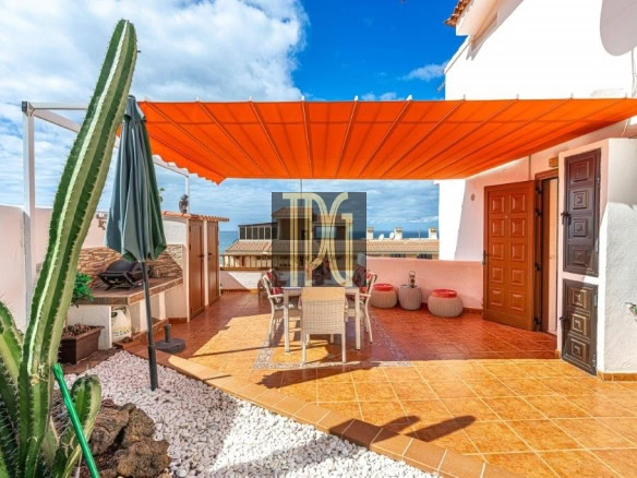 Outdoor rooftop terrace with an orange shade canopy, dining table and chairs, terracotta tiles, and a tall cactus on the left, ocean beyond.