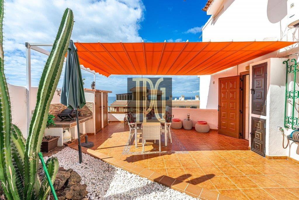 Outdoor rooftop terrace with an orange shade canopy, dining table and chairs, terracotta tiles, and a tall cactus on the left, ocean beyond.