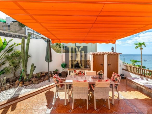 Sunlit seaside terrace with an orange canopy, dining table and wicker chairs, and a view of the sea.