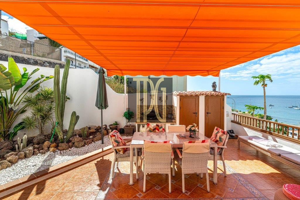 Sunlit seaside terrace with an orange canopy, dining table and wicker chairs, and a view of the sea.