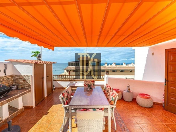 Rooftop terrace with an orange awning, outdoor dining table and chairs, and an ocean view beyond buildings and a grill on the left.