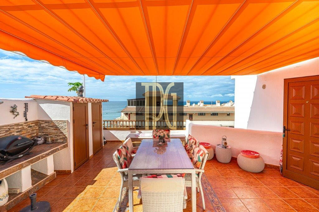 Rooftop terrace with an orange awning, outdoor dining table and chairs, and an ocean view beyond buildings and a grill on the left.