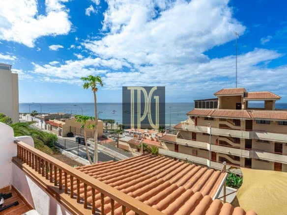 Terracotta-tiled balcony railing overlooking a coastal town with palm trees and the sea under a blue sky.