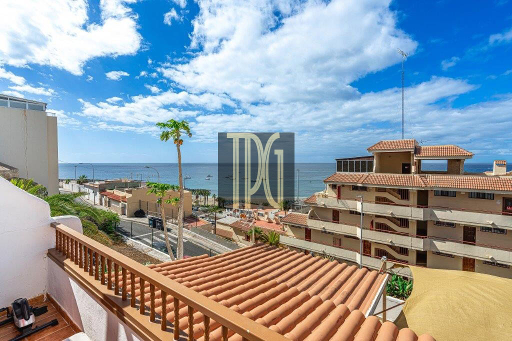 Terracotta-tiled balcony railing overlooking a coastal town with palm trees and the sea under a blue sky.