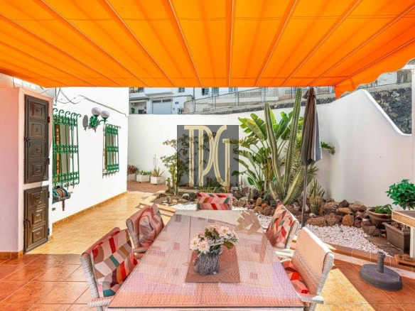 Outdoor dining area under a bright orange canopy, with a long woven table and colorful cushioned chairs on terracotta tiles.