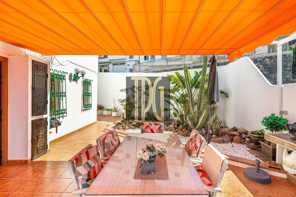 Outdoor dining area under a bright orange canopy, with a long woven table and colorful cushioned chairs on terracotta tiles.