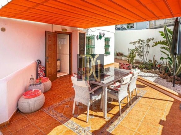 Outdoor patio with an orange sunshade over a glass-top dining table and white wicker chairs on terracotta tiles, plants along a white wall.