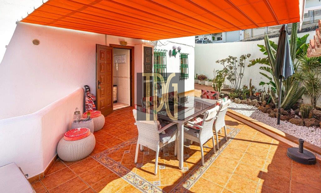 Outdoor patio with an orange sunshade over a glass-top dining table and white wicker chairs on terracotta tiles, plants along a white wall.