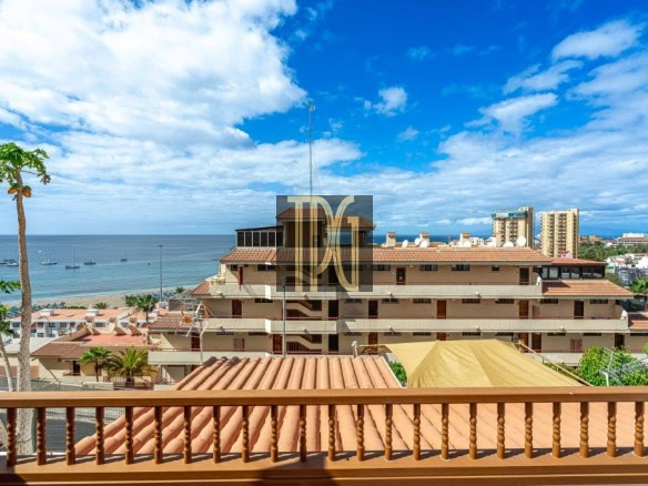 Ocean view from a balcony with a wooden railing, palm trees, terracotta roofs, and a blue horizon of the sea and sky to the left and center.