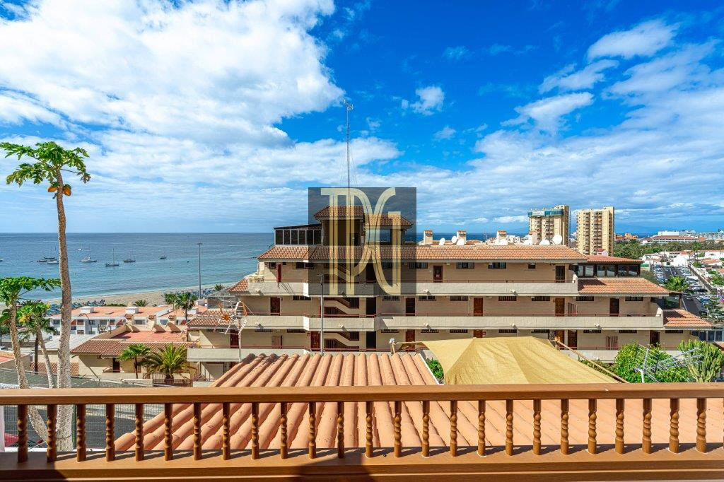 Ocean view from a balcony with a wooden railing, palm trees, terracotta roofs, and a blue horizon of the sea and sky to the left and center.