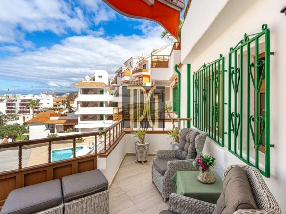 Balcony with wicker seating, a small table with flowers, and green window grilles, overlooking a sunny residential area and pool.”,