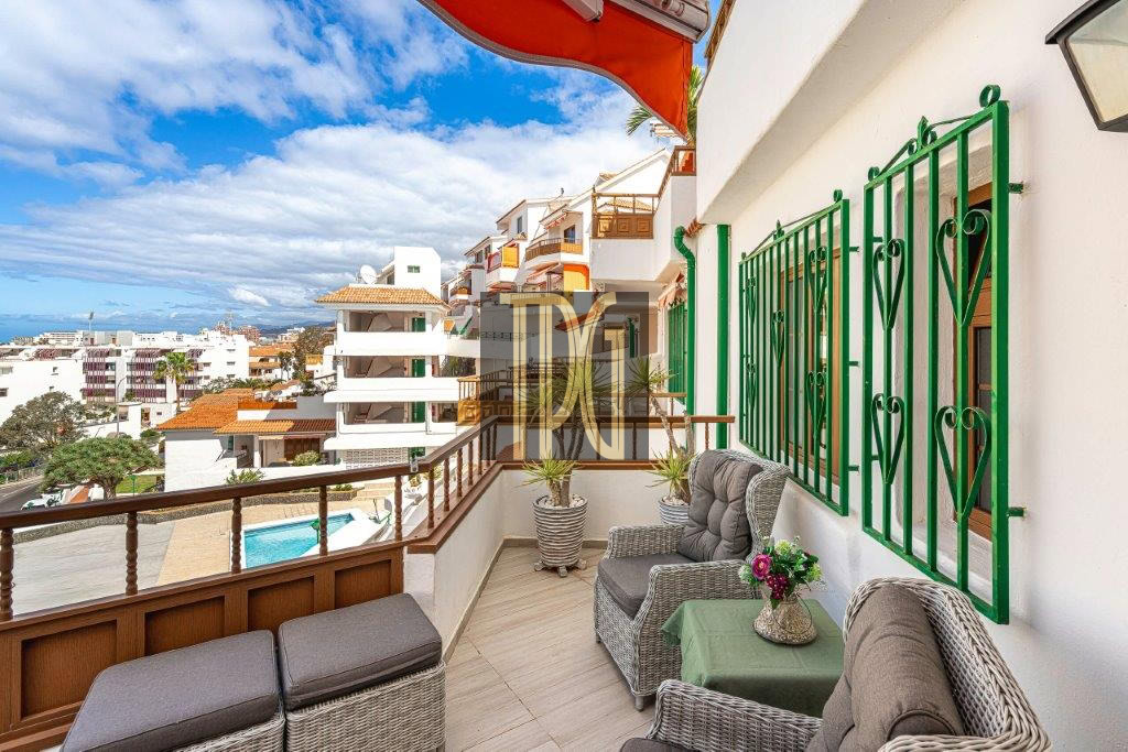 Balcony with wicker seating, a small table with flowers, and green window grilles, overlooking a sunny residential area and pool.”,