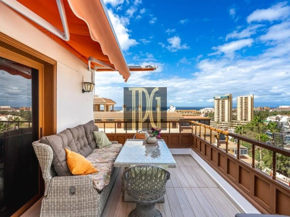 Cozy balcony seating area on an apartment with a glass table, wicker sofa, orange cushions, and an orange awning overlooking the city skyline.