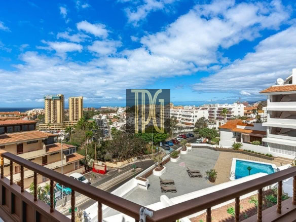 View from a balcony overlooking a seaside town: a pool deck in the foreground, white buildings with terraces, and the ocean on the horizon under a blue sky with clouds.