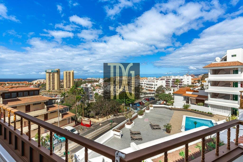 View from a balcony overlooking a seaside town: a pool deck in the foreground, white buildings with terraces, and the ocean on the horizon under a blue sky with clouds.