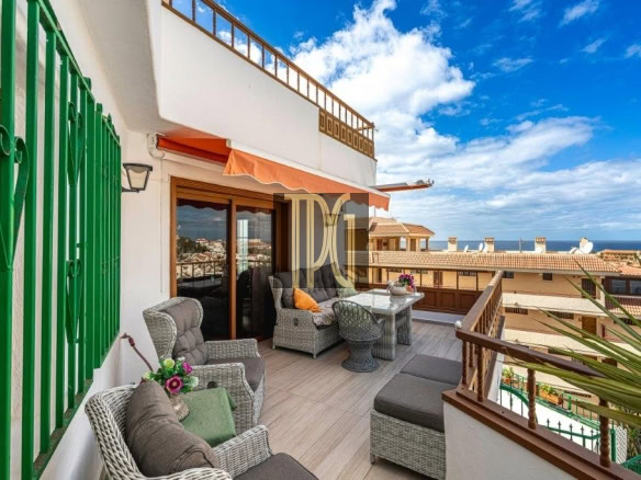 Terrace with wicker seating, glass dining table, and orange awning, overlooking rooftops and the sea on a sunny day.
