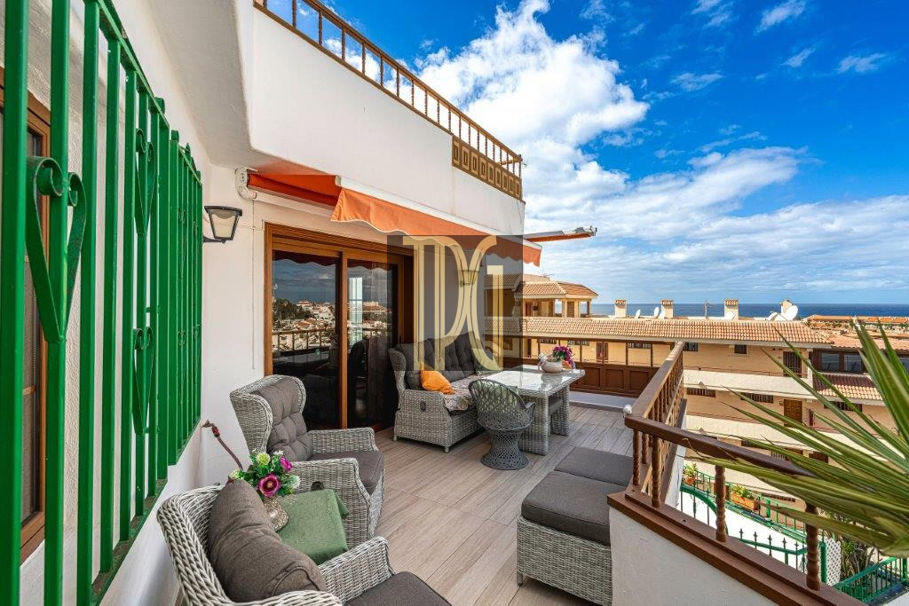 Terrace with wicker seating, glass dining table, and orange awning, overlooking rooftops and the sea on a sunny day.