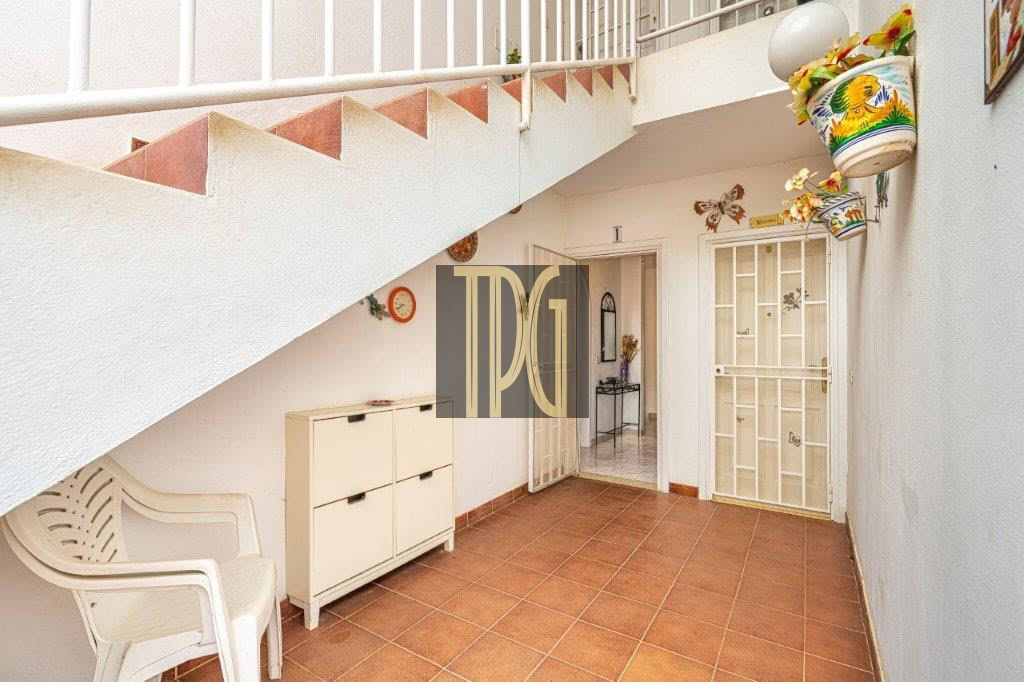 Covered patio with terracotta tiles, white walls, a plastic chair, a cabinet, and glass doors opening to an interior room, under a stairwell.