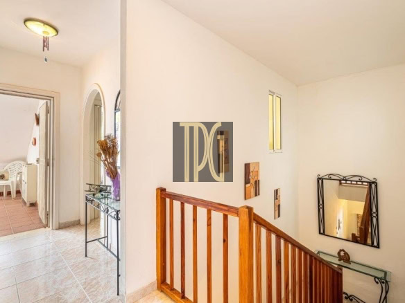 Bright hallway with a wooden staircase railing, a decorative wall mirror, and a glass console table by an arched doorway to a room with white chairs nearby.