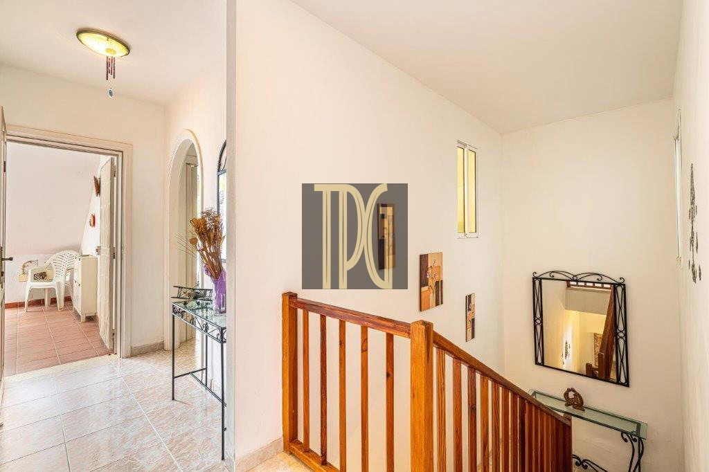 Bright hallway with a wooden staircase railing, a decorative wall mirror, and a glass console table by an arched doorway to a room with white chairs nearby.