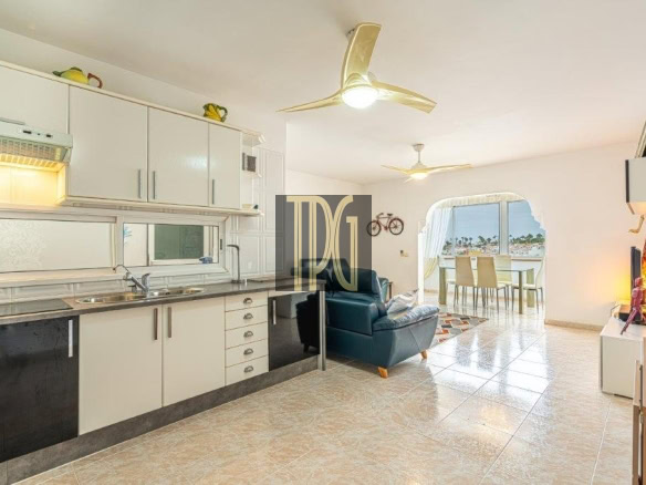 Open-plan living area with white kitchen cabinets, a teal armchair, and a balcony with a table overlooking the sea.