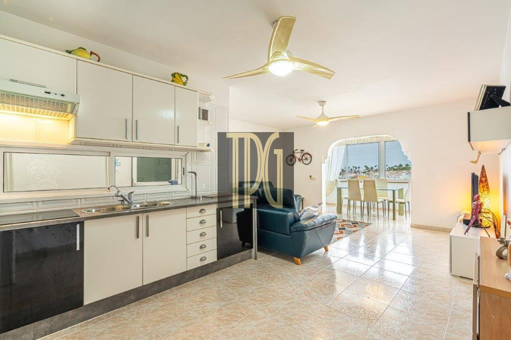 Open-plan living area with white kitchen cabinets, a teal armchair, and a balcony with a table overlooking the sea.
