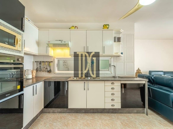 Open-plan kitchen with white cabinets, built-in appliances, and a teal living room sofa visible to the right.