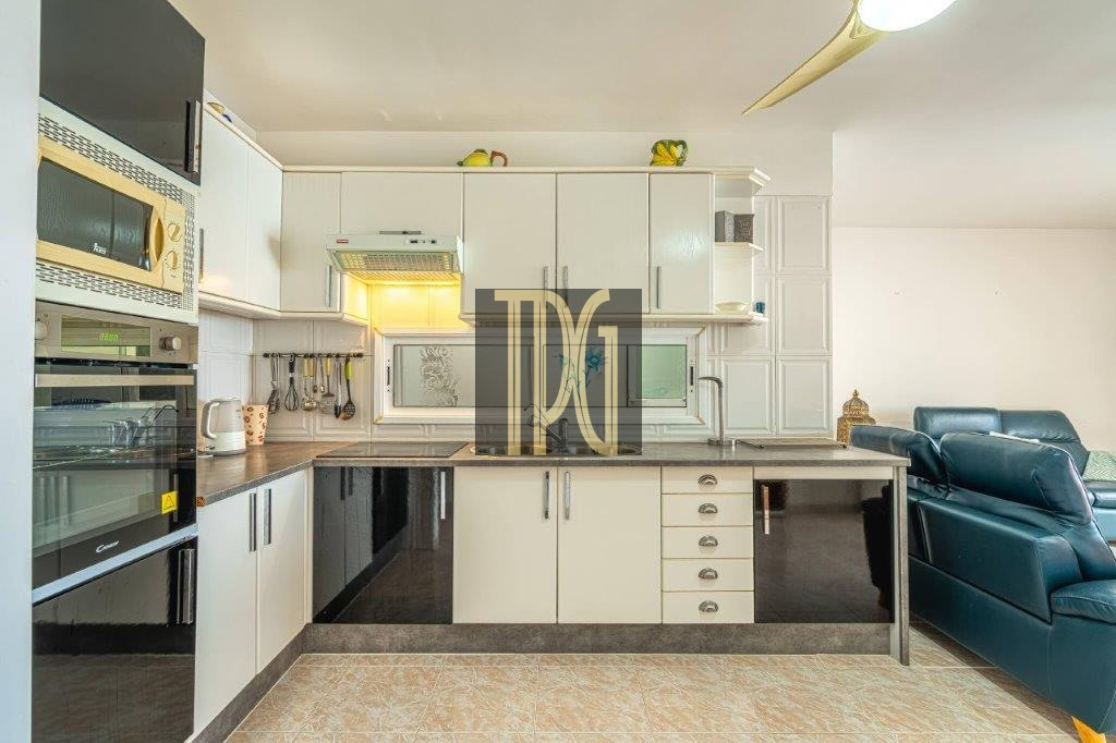 Open-plan kitchen with white cabinets, built-in appliances, and a teal living room sofa visible to the right.