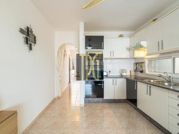 Bright L-shaped kitchen with white cabinets, stainless steel sink, and tiled floor. A hallway with an archway is visible in the background.