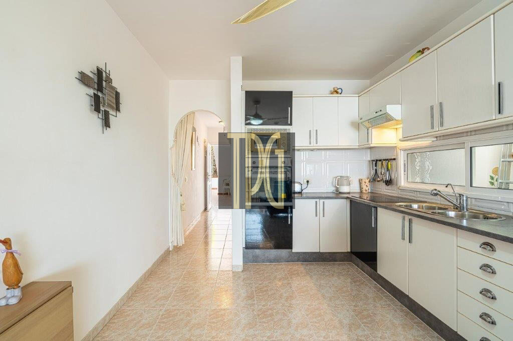 Bright L-shaped kitchen with white cabinets, stainless steel sink, and tiled floor. A hallway with an archway is visible in the background.
