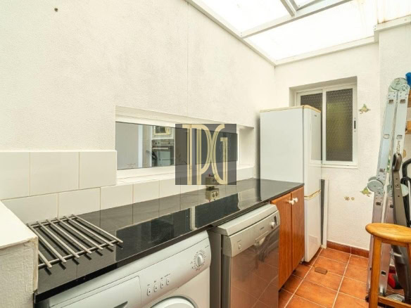 Bright laundry room with a front-loading washer, a dryer, and a white fridge along a black countertop; a ladder and wooden stool rest in the corner under a skylight.