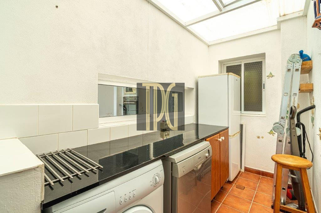 Bright laundry room with a front-loading washer, a dryer, and a white fridge along a black countertop; a ladder and wooden stool rest in the corner under a skylight.
