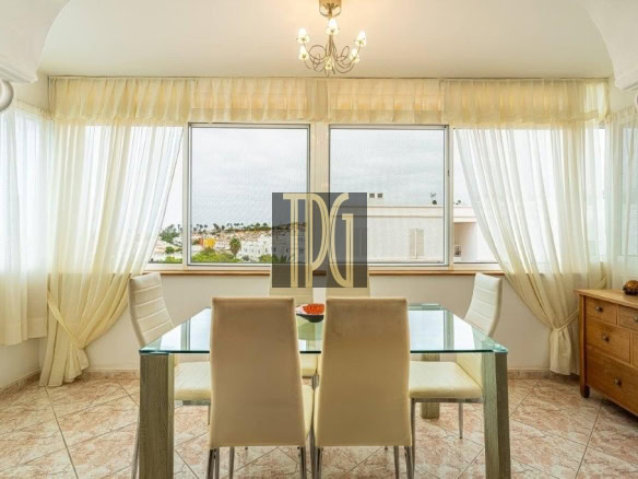 Sunlit dining room with a glass table and cream chairs, sheer curtains, and a chandelier, overlooking a city view through large windows.