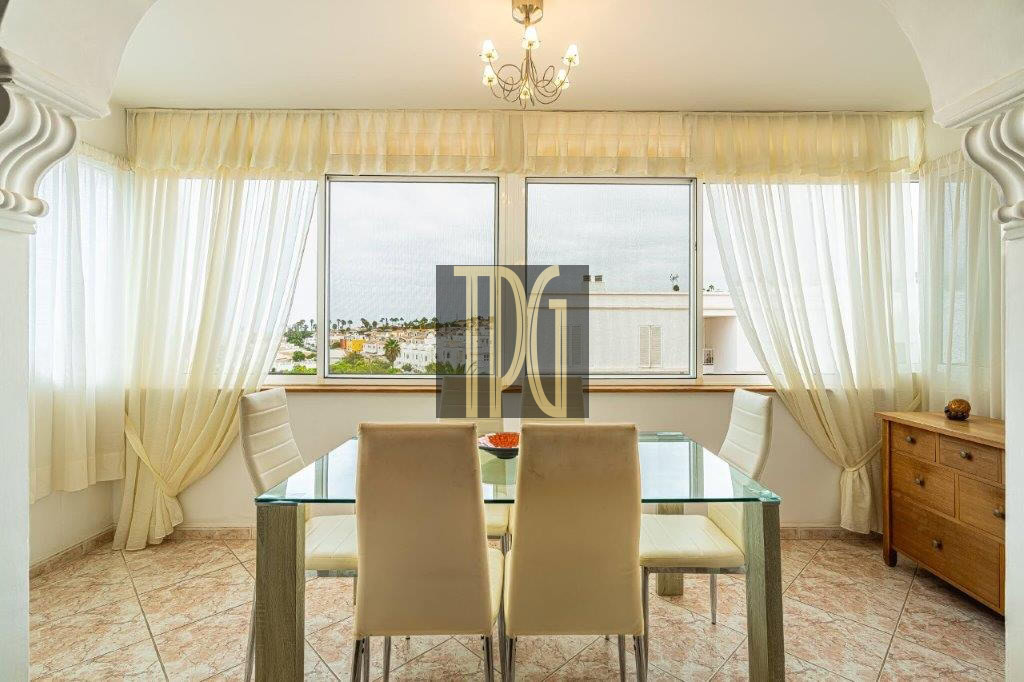 Sunlit dining room with a glass table and cream chairs, sheer curtains, and a chandelier, overlooking a city view through large windows.