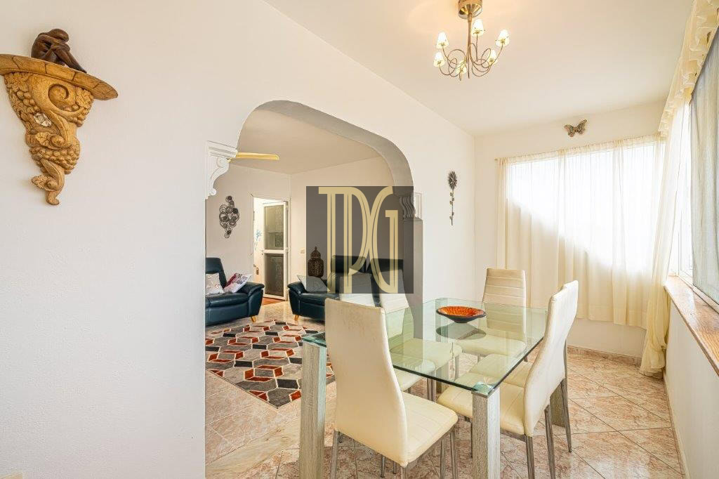 Bright dining area with a glass table and cream chairs, archway to a living room with blue seating and geometric rug—sunlit through sheer curtains.