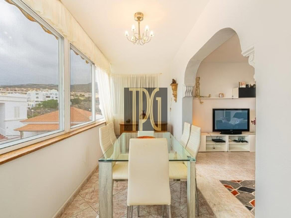 Bright dining area with glass table and white chairs by large windows overlooking rooftops.