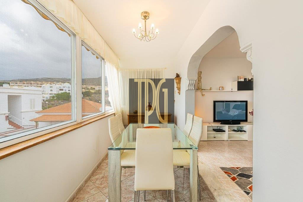 Bright dining area with glass table and white chairs by large windows overlooking rooftops.