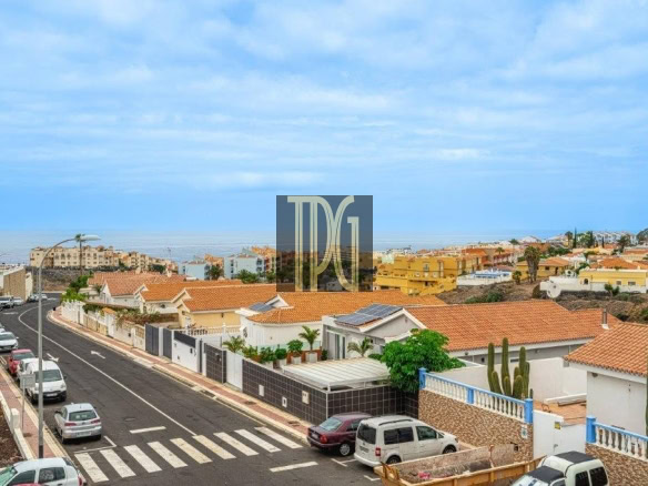 Suburban street lined with white and tan houses with terracotta roofs, the sea on the horizon beyond.