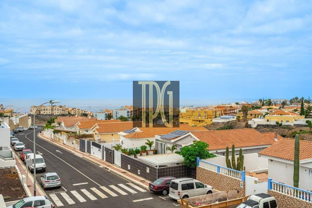 Suburban street lined with white and tan houses with terracotta roofs, the sea on the horizon beyond.