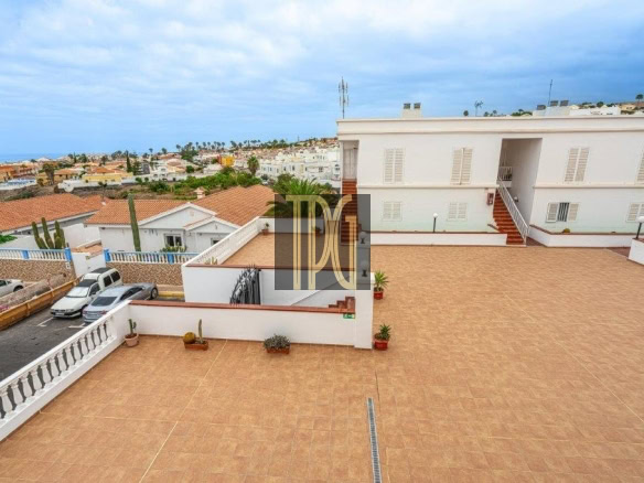 Spacious rooftop terrace with terracotta tiles, white building, stairs, and several potted plants overlooking a coastal town.