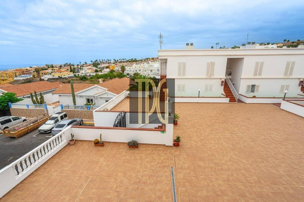 Spacious rooftop terrace with terracotta tiles, white building, stairs, and several potted plants overlooking a coastal town.