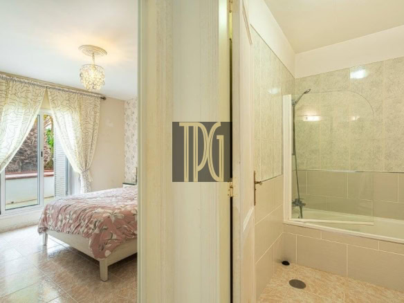 Bright bedroom with a pink floral bedspread, sheer curtains, and a balcony door on the left, adjacent to a tiled bathroom with a bathtub on the right